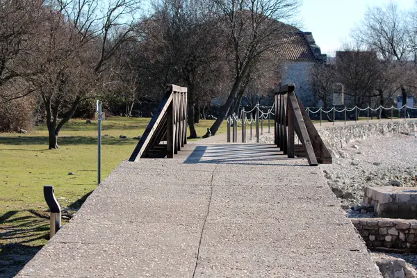 Koyu ahşap korkulukları ve çitleri olan sağlam beton bir iskele taşların üzerindeki çimenli bir sahilden kış güneşinde yapraksız ağaçların gölgelediği eski bir binaya doğru uzanıyor.