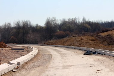 Dolambaçlı, bitmemiş bir yol çıplak toprak ve alçak tepelerden oluşan bir manzarayı ikiye ayırıyor. Yoğun yapraksız ormanlara doğru gidiyor. Yarı doğal bir ortamda sivil çalışmaların ilerlediğini vurguluyor.