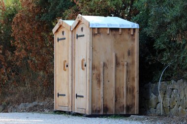 Birbirine bitişik iki ahşap ek tuvalette, orman kenarındaki çakıl taşları üzerinde kırsal veya kamp temizliği için ihtiyaç duyulan, yüksek çatılı çatılar ve ayırt edici kalp kapakları yer alıyor.