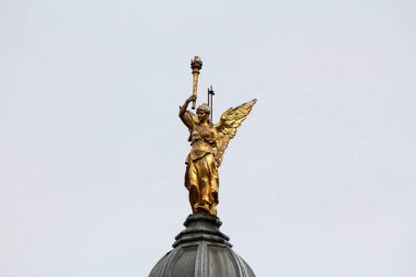 Kanatlı bir kadın heykelinin meşale tutan yaldızlı heykeli, açık gökyüzüne karşı zafer ve ihtişamı temsil eden bir binanın kubbesinde belirgin bir şekilde duruyor.