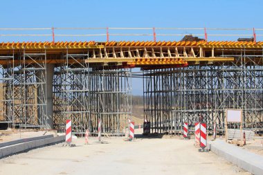 Montaj altındaki büyük bir köprü, yoğun yapı iskelesi ve biçimlendirmeyle desteklenen ve tamamlanmamış alt geçit ve faal çalışma alanını gösteren uyarı işaretleriyle bir inşaat alanını kapsar.