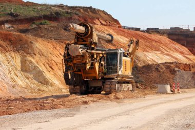 Büyük bir döner sondaj kulesi toprak bir yolda çok renkli bir kazı alanında, tekerlekleri, toprak lekeli metali ve ağır mekanik bileşenleri sergiliyor.