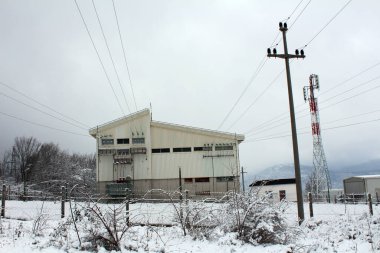 Karlı çitlerle ve yapraksız ağaçlarla çevrili, üstten elektrik hatları ve kafes telekom kulesiyle kışın bir dağ zeminine karşı metal kaplı büyük bir kamu hizmeti istasyonu.