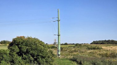 Uzun yeşil metal elektrik direği çimenli tarlalar boyunca elektrik kabloları taşır ve yemyeşil bitki örtüsü, tarım arazileri, evler ve mavi yaz gökyüzü.