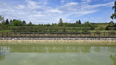 Taş ve ahşap set çizgileri gölün kenarında, arkasında yeşil çalılar ve parlak gün ışığında yakalanan durgun suda ayna benzeri yansımalar var.