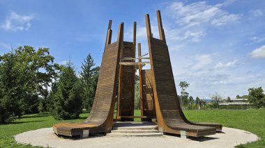 Çağdaş, eğri bankları ve dikey kirişleri olan eşsiz ahşap yapı beton temelde duruyor, kentsel park ortamında yemyeşil ve mavi gökyüzü ile çevrili.