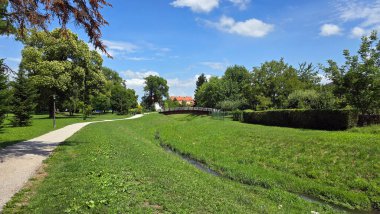 Küçük bir yaya köprüsü ve dolambaçlı park yolları olan yumuşak bir dere bereketli yeşil çimenler, olgun ağaçlar ve mavi gökyüzü tarafından çerçevelenir ve yazın manzaralı bir gezintiye davet edilir.
