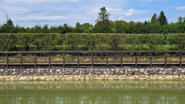 Güvenli çitli düz ahşap geçit yeşil çitlere paralel uzanır, taş zemine kurulur ve güneşli bir park göletinin durgun yüzeyine yansıtılır.
