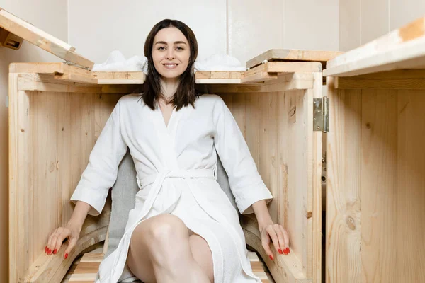 A young European brunette girl in a white robe is sitting in a cedar barrel, smiling and looking at the camera. Salon procedures. High quality photo