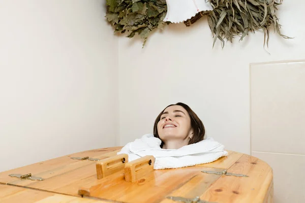 Satisfied young European brunette in a cedar barrel, smiling with closed eyes. Salon procedures. High quality photo