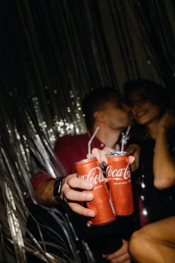 CHERNIGIV, UKRAINE - 21 Kasım 2022: Genç bir çift gri yağmur perdesine karşı Coca-Cola tutarken yerde oturup öpüşüyor. Yüksek kalite fotoğraf