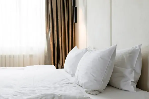 在明亮的现代酒店房间里 有一张豪华整洁的床 上面铺着毛绒绒的白色枕头 柔和的灯光凸显了布景的典雅与舒适 — 图库照片