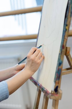 Bir ressamın yakın plan resmi. Elleri ustalıkla tuvale çizim yapıyor. Parlak bir sanat stüdyosundaki renkli bir sehpaya konur..