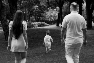 Bir ailenin yemyeşil bir parkta yürürken huzurlu bir anını yakalayan siyah beyaz bir görüntü. Çocuk, kalın gövdeli ağaçlarla çevrili çimenli bir yolda yetişkinlerin önünde yürüyor.