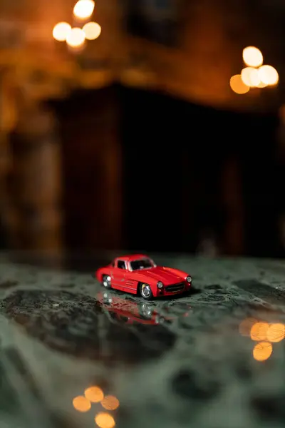 A vibrant red toy car on a textured surface, illuminated by soft bokeh lights in the dark background.