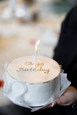 Bir insan güzel bir doğum günü pastası tutar, zarif altın harflerle süslenir ve bir mum yakar, kutlamanın sıcaklığını ve neşesini yakalar.