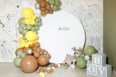 Beyaz dairesel bir arkaplanı çevreleyen renkli balonlar ilk doğum günü kutlamasını simgeleyen yazılı bir arkaplan. Bir çizgi film köpeği ve heceleme çocuğu bloklar Neşeli bir dokunuş ekleyin