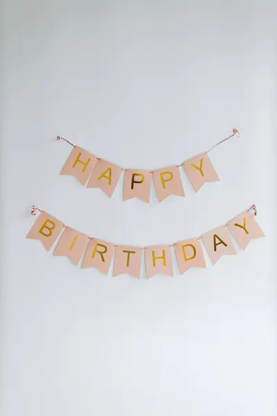 stock image A tasteful Happy Birthday banner, with each letter printed on separate pastel pink flags, is strung in two neat rows against a pristine white wall, exuding warmth and celebration