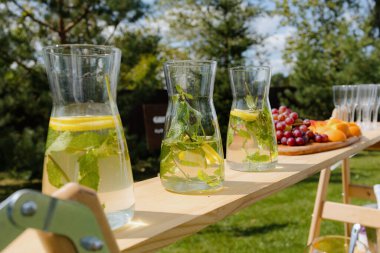 Limon ve limon dilimleri, nane yaprakları ve güneşli bir günde taze üzüm ve portakalların eşlik ettiği cam sürahilerin yer aldığı bir açık hava içecek istasyonu..