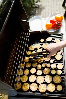 Slices of eggplant and colorful bell peppers are being grilled outdoors as sunlight illuminates the backyard. Tongs are used to turn the vegetables, showcasing vibrant colors and textures. clipart