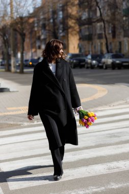 Şık, siyah paltolu bir kadın, şehirde güneşli bir günün tadını çıkarırken, canlı bir lale buketini tutarak yaya geçidinde yürüyor, park etmiş arabalar ve şehir manzarasıyla çevrili..
