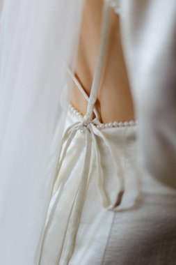 Yumuşak bir elbisenin yakın görüntüsü zarif kurdelelerle bağlanmış bir yayı sergiliyor, zarif bir yaz tarzını yansıtıyor. Açık renk kumaş zarif bir şekilde dış mekanın sıcak zeminine çarpar..