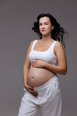 Işıl ışıl hamile bir anne, kendinden emin bir şekilde durur ve bebeğinin karnını doğal bir zarafetle nazikçe kucaklar. Yumuşak ışıklandırma, evin içinde huzurlu bir ortamda neşe dolu beklentisinin altını çiziyor..