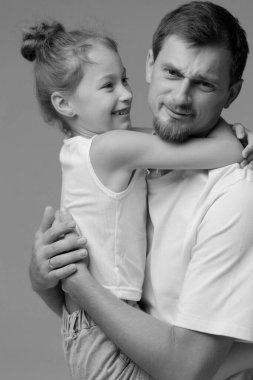 Neşeli küçük bir kız babasını kucaklarken o da neşeli ve abartılı bir yüz ifadesi takınıyor. Bağları sevgi ve mutluluk saçıyor, aralarındaki değerli bağı minimalist bir ortamda gösteriyor..