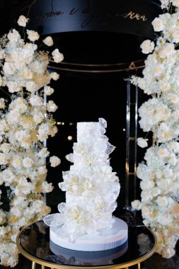 Göz kamaştırıcı bir düğün pastası zarif bir şekilde yuvarlak bir masanın üzerinde duruyor. Etrafı bembeyaz çiçeklerle çevrili. Arkaplan ışığın altında parıldıyor ve akşam için romantik bir ortam yaratıyor..