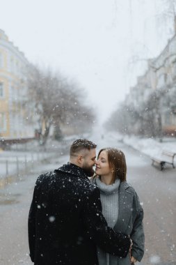 İki aşık, karlı bir caddenin ortasında yumuşak kar taneleriyle çevrili bir öpüşmeyi paylaşıyor. Sıcak atmosfer bu kış günündeki sevgilerini vurguluyor..