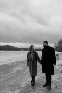 İki insan, sıcak paltolara sarılı, karlı bir nehir kıyısında el ele yürüyor. Kahkahaları kışın serin havasıyla karışıyor. Bulutlu gökyüzü bu huzurlu ana duygusal bir hava katıyor..