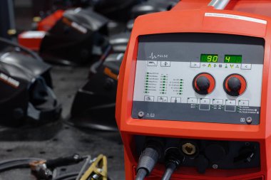 Kaynak makinesinin canlı bir görüntüsü ön planda duruyor, miğferler ve aletlerle çevrili, yoğun bir atölyenin özünü yakalıyor..