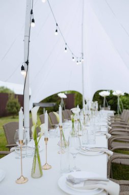 Beyaz çarşaflı açık hava düğün masası, altın mum tutacakları, ve beyaz bir çadırın altında sicim ışıkları olan kalla zambağı vazoları.