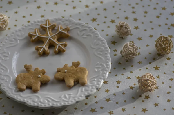 Painted traditional Christmas gingerbreads arranged on white plate, tablecloth with golden stars, snowflake and reindeer shapes with ball arrangements