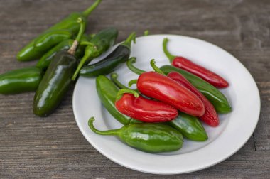 Capsicum annuum Jalapeno acı biber hasadı, yeşil ve kırmızı meyveler beyaz tabakta, ahşap kahverengi masada.