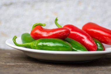 Capsicum annuum Jalapeno acı biber hasadı, yeşil ve kırmızı meyveler beyaz tabakta, ahşap kahverengi masada.