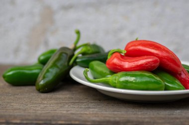 Capsicum annuum Jalapeno acı biber hasadı, yeşil ve kırmızı meyveler beyaz tabakta, ahşap kahverengi masada.
