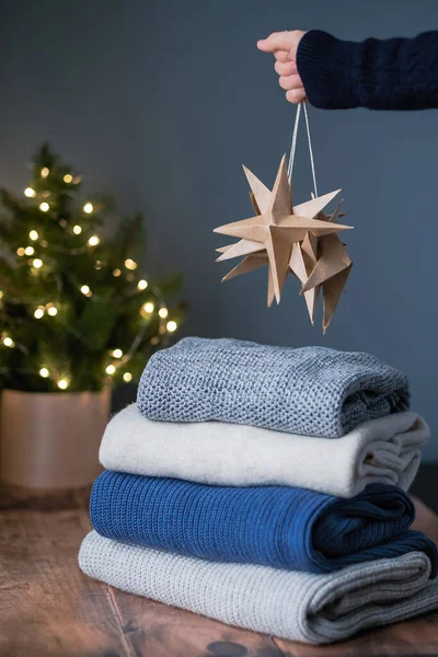 A stack of knitted sweaters with sustainable Christmas decoration on the background and a Christmas tree with lights. Holiday online shopping. Sustainable fashion. High quality photo