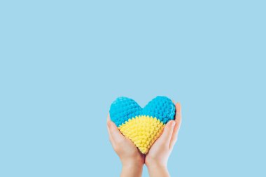 Handmade crochet amigurumi heart in the hands of a child on yellow and blue colors. Flag of Ukraine. Isolated on a blue background. Colors of the flag of Ukraine. Space for text. High quality photo
