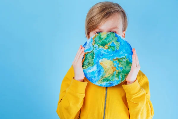 A child closing his face with a painting planet Earth, eyes looking at the camera on a blue background. Save go earth, saving environment, save clean planet, ecology concept. Sustainability project