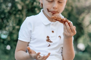 Çikolatalı pasta yiyen bir çocuk parktaki ahşap bankta oturuyor, yüzü ve eli erimiş çikolata içinde. Beyaz elbiselerde kirli çikolata lekeleri. Açık havada. Yüksek kalite fotoğraf