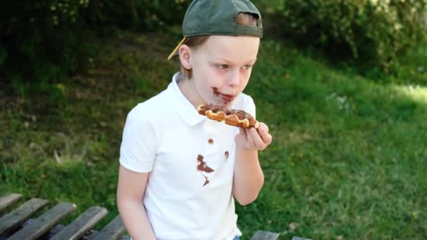 一个小孩坐在公园里的木制长椅上吃着一块巧克力蛋糕 脸朝天 交上融化的巧克力 白色衣服上有肮脏的巧克力污渍 高质量的照片 — 图库视频影像