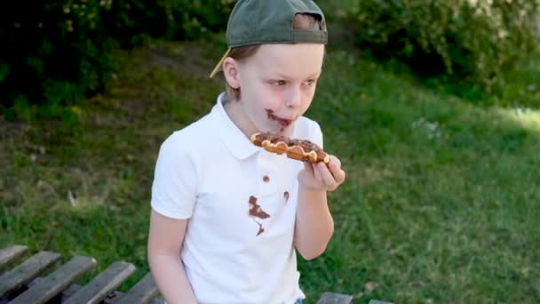 公園の木のベンチに座ってチョコレートケーキを食べている子供は 顔と溶かしたチョコレートで手 白い服に汚れたチョコレートの汚れ 屋外で — ストック動画