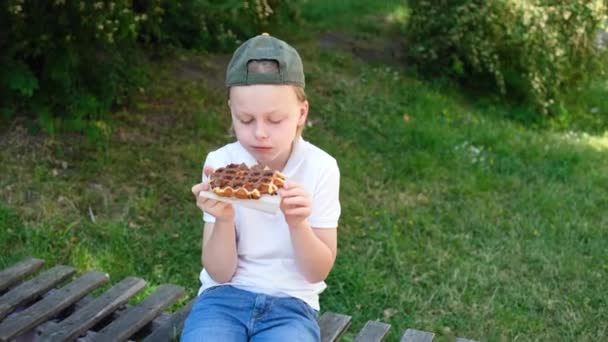 一个小孩坐在公园的木制长椅上吃巧克力蛋糕 — 图库视频影像