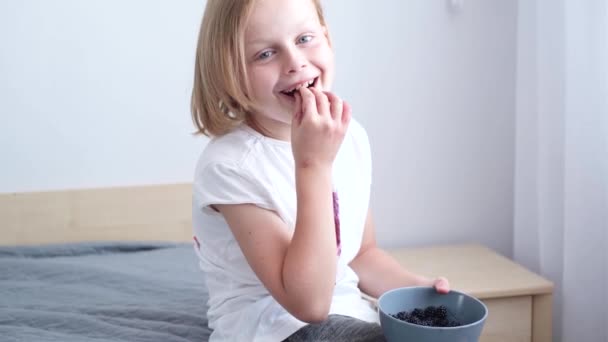 肖像画的孩子正在吃浆果 坐在房间的床上 白色衣服上肮脏的蓝莓色 日常生活污迹的概念 高质量的4K镜头 — 图库视频影像