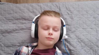Beyaz kulaklıklı genç yakışıklı bir çocuk bir ev yatağında yatıyor. Müzikle rahatla. Müzik dinler ve evde şarkılar söyler. Üst Manzara. Yüksek kalite 4k görüntü