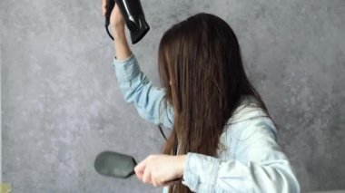 Kız gri arka planda saç fırçası kullanarak saç kurutma makinesiyle ıslak saçı kurutuyor. Saçını iyi taramışsın. Yüksek kalite 4k görüntü