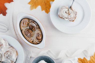 Freshly baked cinnamon rolls sit in a dish beside sliced apples, a glass of milk, and a bowl of ice cream, surrounded by autumn leaves. High quality photo