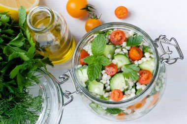 Cam kavanozda taze kuskus salatası, salatalık, domates, otlar, limon ve beyaz bir zemin. Tabbouleh salatası. Sağlıklı vejetaryen yemekleri. Üst Manzara. Yüksek kalite fotoğraf