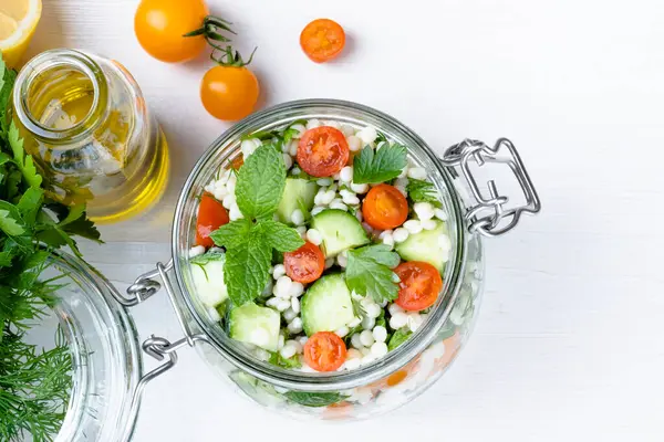 Cam kavanozda taze kuskus salatası, salatalık, domates, otlar, limon ve beyaz bir zemin. Tabbouleh salatası. Sağlıklı vejetaryen yemekleri. Üst Manzara. Yüksek kalite fotoğraf